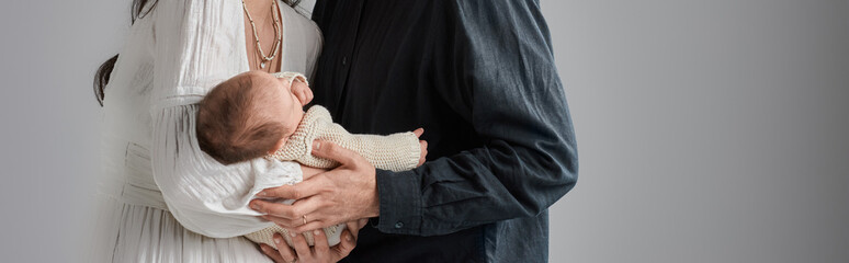 adorable newborn baby boy in arms of his loving parents on gray backdrop, family concept, banner