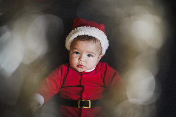 exciting smiling baby boy in red santa claus costume with christmas hat unwrapping christmas gifts and plays with balls at home on the floor in the living room