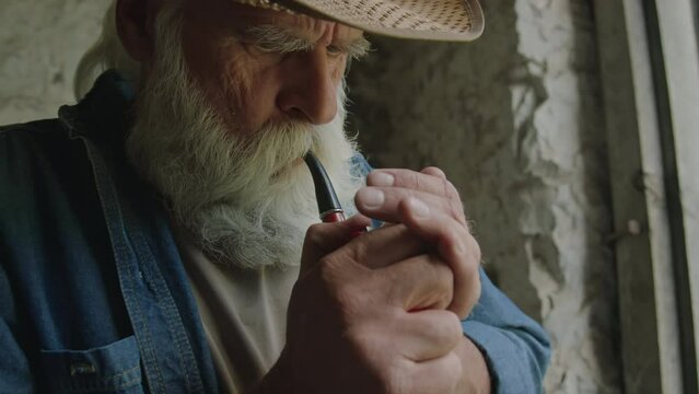 Close up of aged bearded man wearing hat smoking pipe while resting indoors