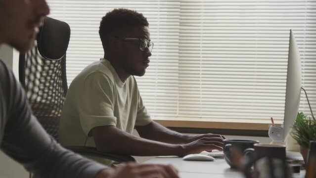 Side view waist up shot of two multiracial male software developers sitting at desk in front of computer monitors in modern open space office