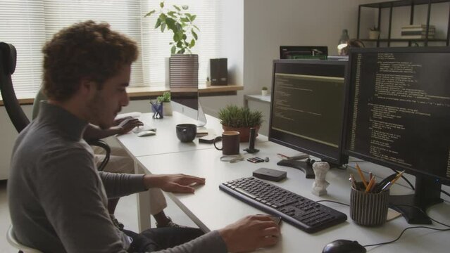 Biracial curly haired handsome guy sitting down at office desk in front of two computer monitors and starting writing program code on keyboard, working with diverse colleagues in office