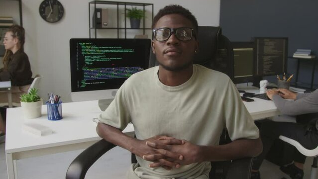 Medium portrait of African American male programmer sitting relaxedly in office chair and looking at camera while working with diverse colleagues in open space office