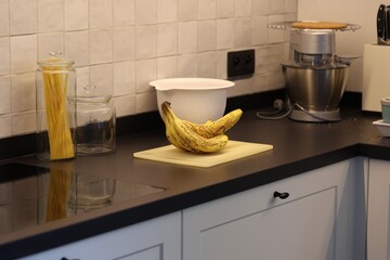 A portrait of a bunch of yellow bananas with brown spots lying on a white plastic cutting board on a black kitchen countertop. The delicious energizing food is ready to eat and ideal before sport.