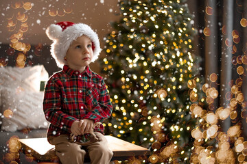 A beautiful little boy in a Santa Claus hat is sitting on a table against the background of a Christmas tree and a beautiful bokeh. The concept of celebrating Christmas and New Year 2024.