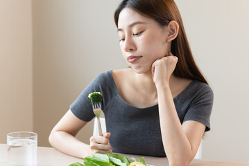 unhappy asian women is on dieting time looking at broccoli on the fork. girl do not want to eat vegetables and dislike taste - obrazy, fototapety, plakaty