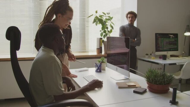 Medium shot of diverse couple of programmers looking at computer monitor and having discussion while their colleagues standing by windowsill and watching at them, working together in open space office