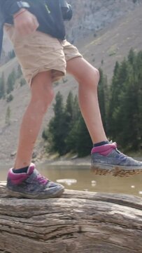 A young adventurer strolls along a fallen tree by a mountain lake in a U.S. national park. Focus on legs and boots, showcasing a childhood immersed in nature. The vertical video captures the essence