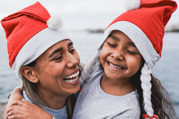Portrait of happy latin mother and child wearing santa claus hat outdoor - Mom and daughter...