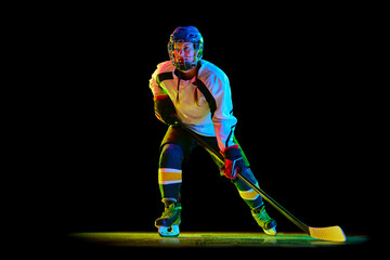 Young girl, hockey player in uniform and helmet training, standing with stick against black studio background in neon light. Concept of professional sport, competition, game, action, hobby