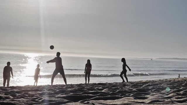 A group of people with children on a beach near the sea playing beach volleyball during sunset in backlight.