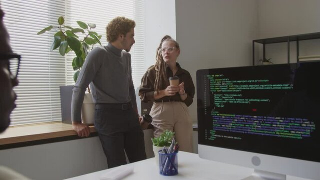 Medium shot of multiethnic couple of programmers talking during coffee break in office while their African American male colleague writing software program code on desktop computer in foreground