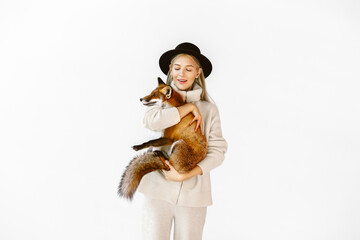 A girl in a white pullover, and black hat  holds a red fox in her hands. The blonde in studio. Pretty woman with fox on white background.