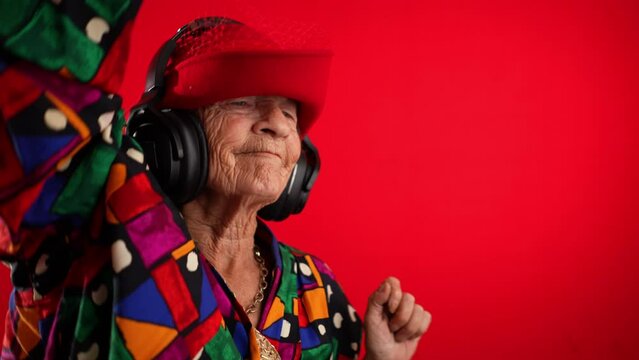 Funny happy old elderly woman with no teeth listening and dancing to music on smart phone in hand with headphones on red background in slow motion
