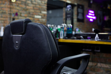 Back of a black leather chair with armrests in a barbershop in front of a mirror and a hairdresser's table with hair care tools