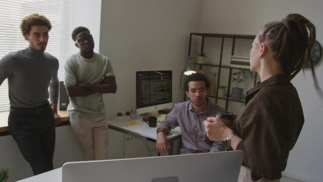 Young enthusiastic Caucasian female programmer with dreadlocks standing beside desktop computer and making software presentation for diverse male team in modern office