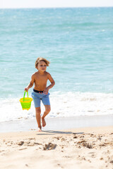 Happy child boy playing with the sand on the beach, pouring water from plastic bucket on the sea beach, building sand castle, enjoying summer. Advertising for tour operators, hotels, airlines.