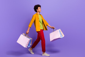 Full length profile side photo of positive man wear stylish clothes carrying packages empty space isolated on purple color background