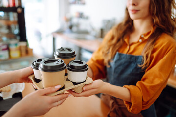 Close-up of a female barista's hands passing prepared drinks to go. A young woman owner of a coffee shop behind the bar prepares coffee. Concept of orders to go.