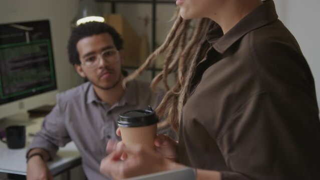 Tilting shot of young creative Caucasian woman with dreadlocks and takeaway paper coffee cup in hand showing presentation of new software to Biracial male programmer in office