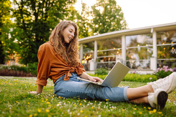 Beautiful woman in casual clothes on a green lawn with a laptop. Happy freelancer woman working on laptop outdoors. Education and freelancing concept.