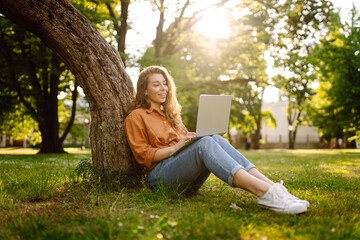 Obraz premium Beautiful woman in casual clothes on a green lawn with a laptop. Happy freelancer woman working on laptop outdoors. Education and freelancing concept.