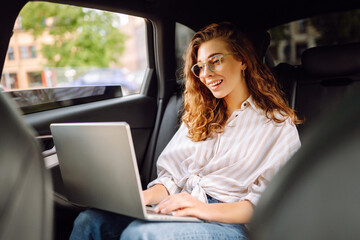 Beautiful business woman in casual clothes with a laptop working in the back seat of a car. Remote work concept, transport, technology.