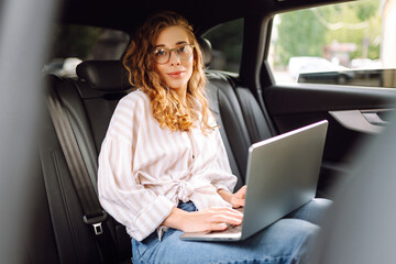 Beautiful business woman in casual clothes with a laptop working in the back seat of a car. Remote work concept, transport, technology.