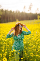 Naklejka na ściany i meble Young woman walking through a flowering field gently touches yellow flowers. A tourist in a straw hat enjoys sunny weather in a rapeseed flowering field. Nature concept.