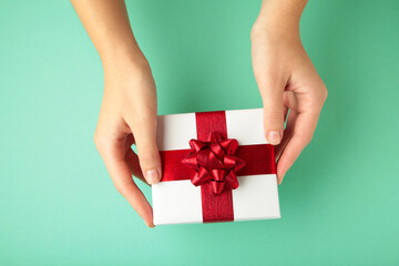 White gift with red bow in female hands on mint background. Space for text.