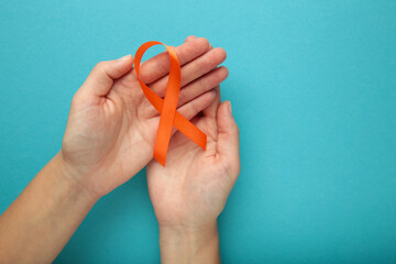 Hands holding orange color ribbon on blue background. Kidney Cancer Awareness, Leukemia disease, Skin cancer awareness, World Cancer Day. - obrazy, fototapety, plakaty