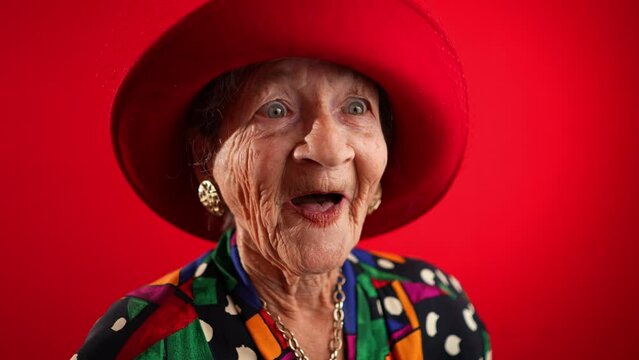 Laughing funny elderly woman with no teeth saying WOW having great success wearing red hat isolated on red background. Studio fisheye portrait caricature in slow motion