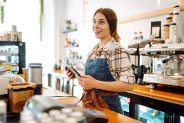 Portrait of a friendly female barista holding a tablet computer and smiling. A young coffee shop owner standing behind the bar using a digital tablet. Takeaway concept, technology.