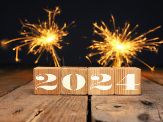 Wooden blocks with the inscription 2024 on a rustic wooden table, fireworks or sparkler in the background