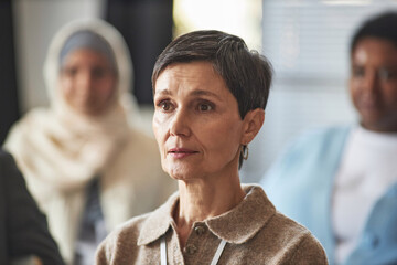 Middle aged brunette woman in casualwear listening to speaker report or presentation while sitting...