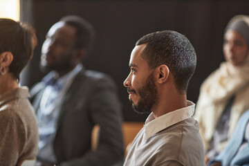 Side view of young bearded multiethnic businessman listening to report of speaker while sitting among intercultural audience at conference - obrazy, fototapety, plakaty