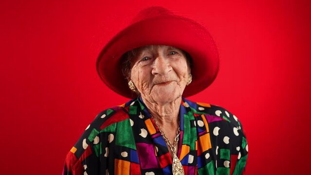 Surprised, happy, funny crazy elderly mature woman with no teeth, 80s, acting shy wearing red hat isolated on red background. Concept of elderly shy person in slow motion.
