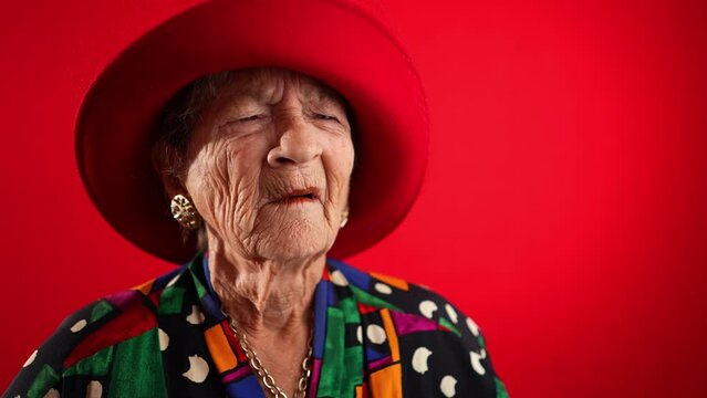 Funny portrait of sick tired headache migraine exhausted elderly mature old woman rubbing temples wearing red hat isolated on red background in studio in slow motion