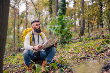 During his woodland hike, a hipster enjoys a rest, sitting on a tree stump while consulting a paper map to plan his route.