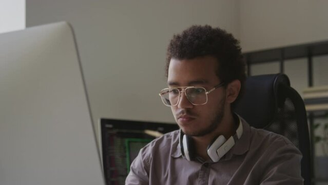 Tilting shot of focused Biracial young man moving eyes over computer screen fast and typing program code on wireless keyboard while working in office