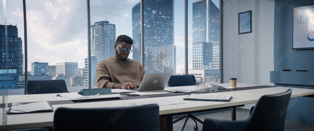 Poster confident businessman sitting at a desk in modern office, using laptop computer next to window with 
