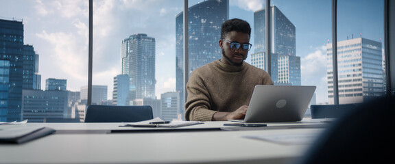 Confident Black Businessman Sitting at Desk in Modern Office, Using Laptop Computer, Next to Window with Big City with Skyscrapers View. Focused Finance Manager Planning Work Projects. - obrazy, fototapety, plakaty