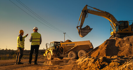 Caucasian Male Real Estate Investor And Engineer Talking On Construction Site Of Apartment Block. Colleagues Discussing Building Progress. Excavator Loading Sand In Industrial Truck On Warm Day © Gorodenkoff