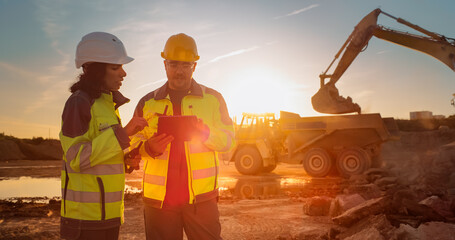 Caucasian Male Engineer Using Tablet And Talking To Hispanic Female Architect On Construction Site Of Real Estate Project. Colleagues Discussing Building Plan Of Apartment Complex During Golden Hour. - obrazy, fototapety, plakaty