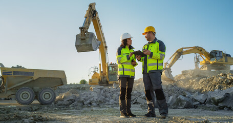Hispanic Female Inspector Talking to Caucasian Male Land Development Manager With Tablet On Construction Site Of Real Estate Project. Industrial Excavators Preparing For Laying Foundation To Building.
