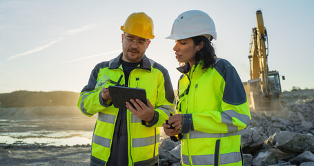 Fototapeta na wymiar Caucasian Male Civil Engineer Talking To Hispanic Female Inspector And Using Tablet On Construction Site of New Apartment Building. Real Estate Developers Discussing Business, Excavators Working.