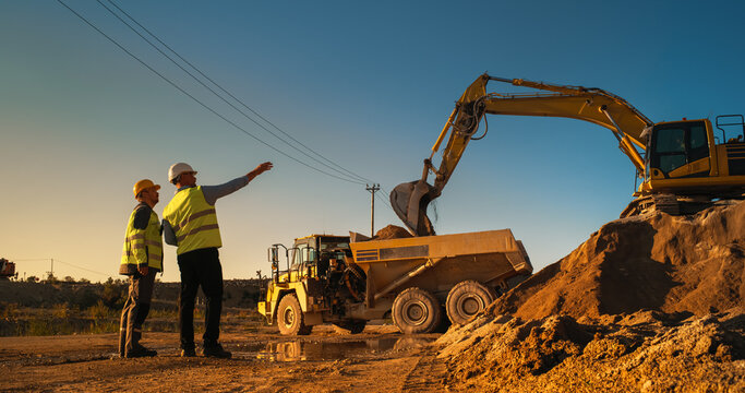 Caucasian Male Real Estate Investor And Engineer Talking On Construction Site Of Apartment Block. Colleagues Discussing Building Progress. Excavator Loading Sand In Industrial Truck On Warm Sunny Day.