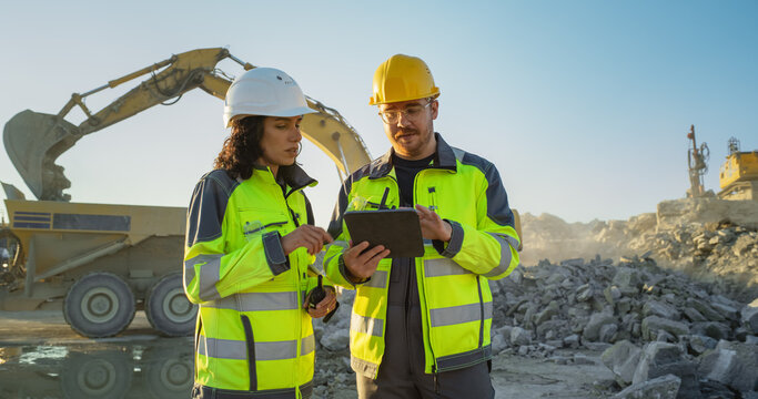 Caucasian Male Civil Engineer Using Tablet And Talking To Hispanic Female Architect On Construction Site Of New Real Estate Project. Diverse Colleagues Discussing Building Plan Of Apartment Block.