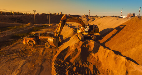Aerial Drone Shot Of Construction Site On Sunny Evening: Industrial Excavator Loading Sand Into A Truck. The Process Of Building New Apartment Block. Workers Operating Heavy Machinery Concept.