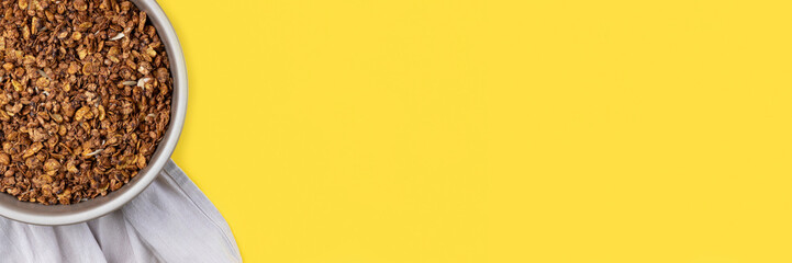 Roasted chocolate granola cereal in round baking tray on bright yellow background banner. Top view, flat lay muesli. Panoramic web header. Wide screen wallpaper