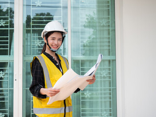 Portrait of woman engineer at building site smiling and looking at camera. Young construction manager standing in yellow safety vest and white hardhat with blueprint paper.